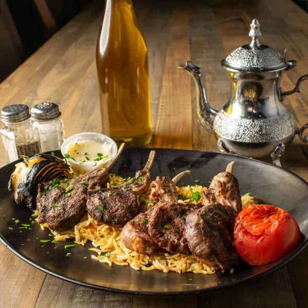 Grilled lamb chops with rice, roasted vegetables, and sauces, alongside an ornate silver teapot.